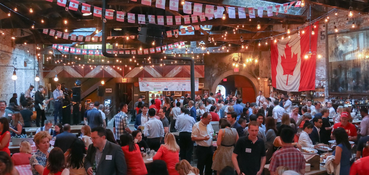 A crowd of people enjoy socializing in a large hall with Canadian flags hanging from the ceiling